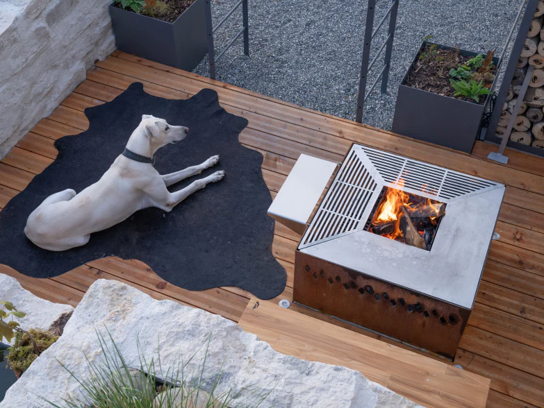 Quadratische Feuerschale mit natürlicher Rostpatina und Grillring auf einer Holzterrasse, daneben liegt ein Hund